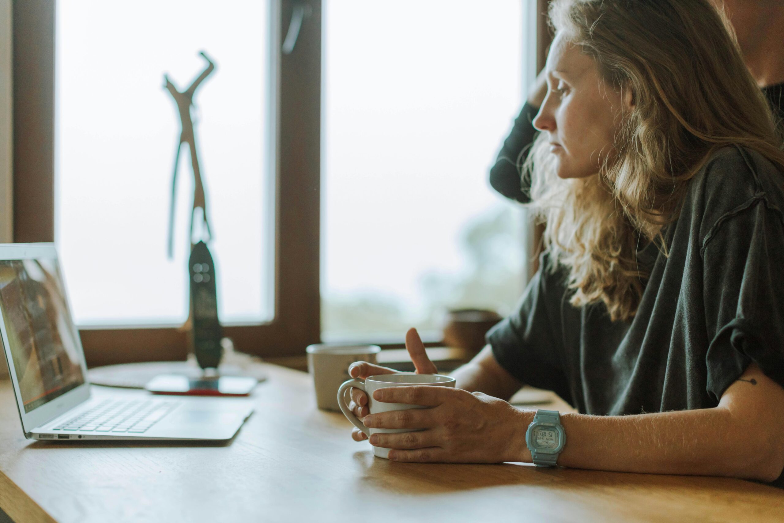 A woman sits by the window, drinking coffee while looking at her laptop in a tranquil indoor setting.