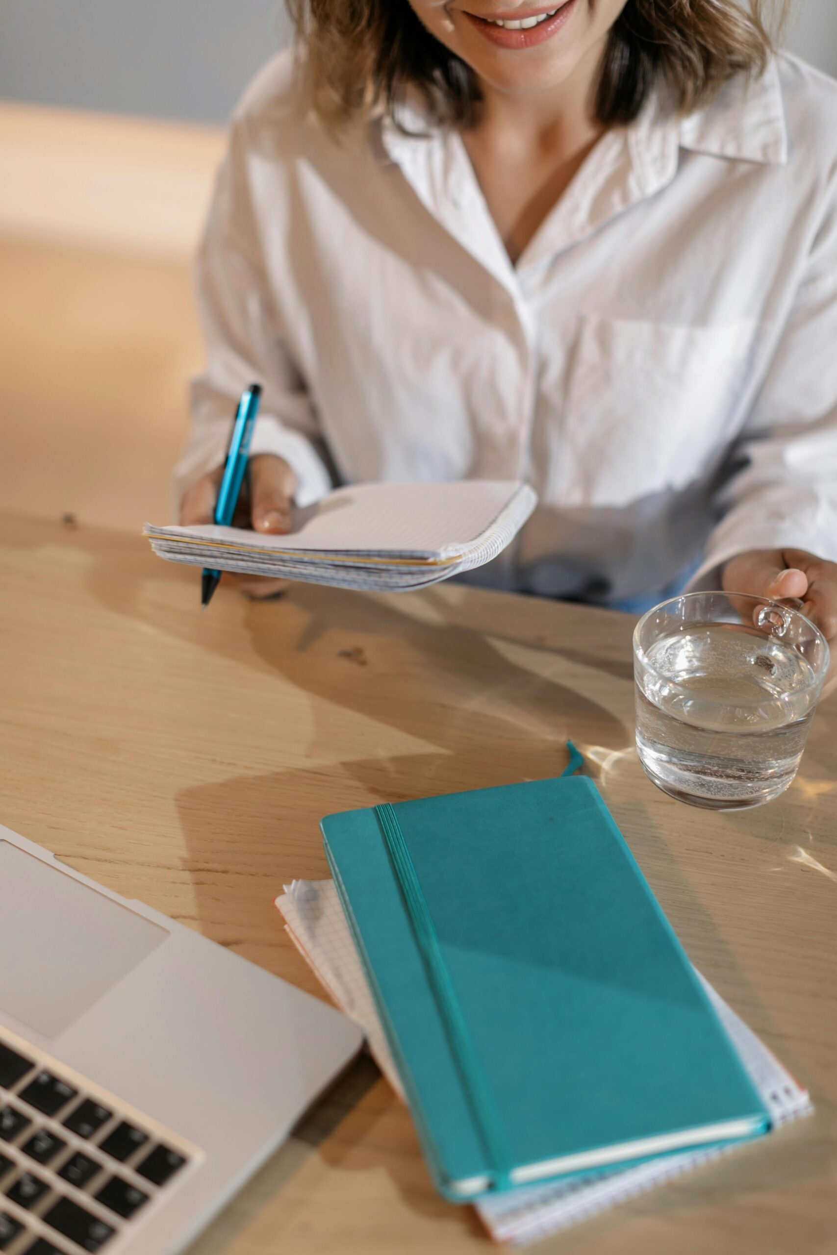 Woman writing notes at a desk with a laptop, notebook, and glass of water.