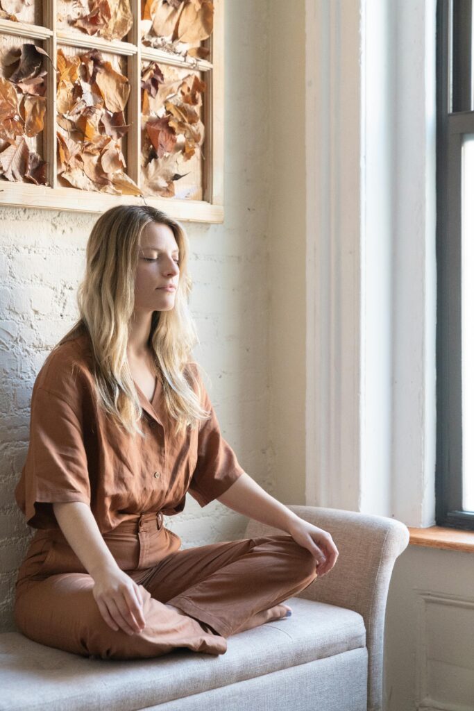 A young caucasian woman sits cross-legged meditating indoors with closed eyes, in a peaceful setting.